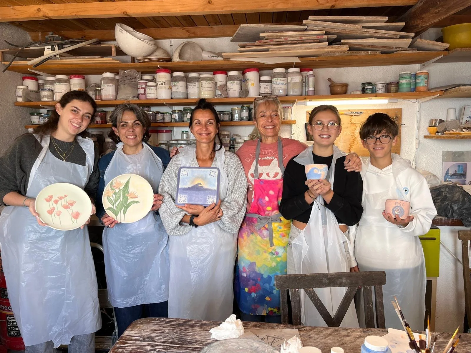 Group of six women and girls in a ceramic painting workshop, holding painted ceramic plates and bowls, in a studio with shelves of pottery supplies behind them.
