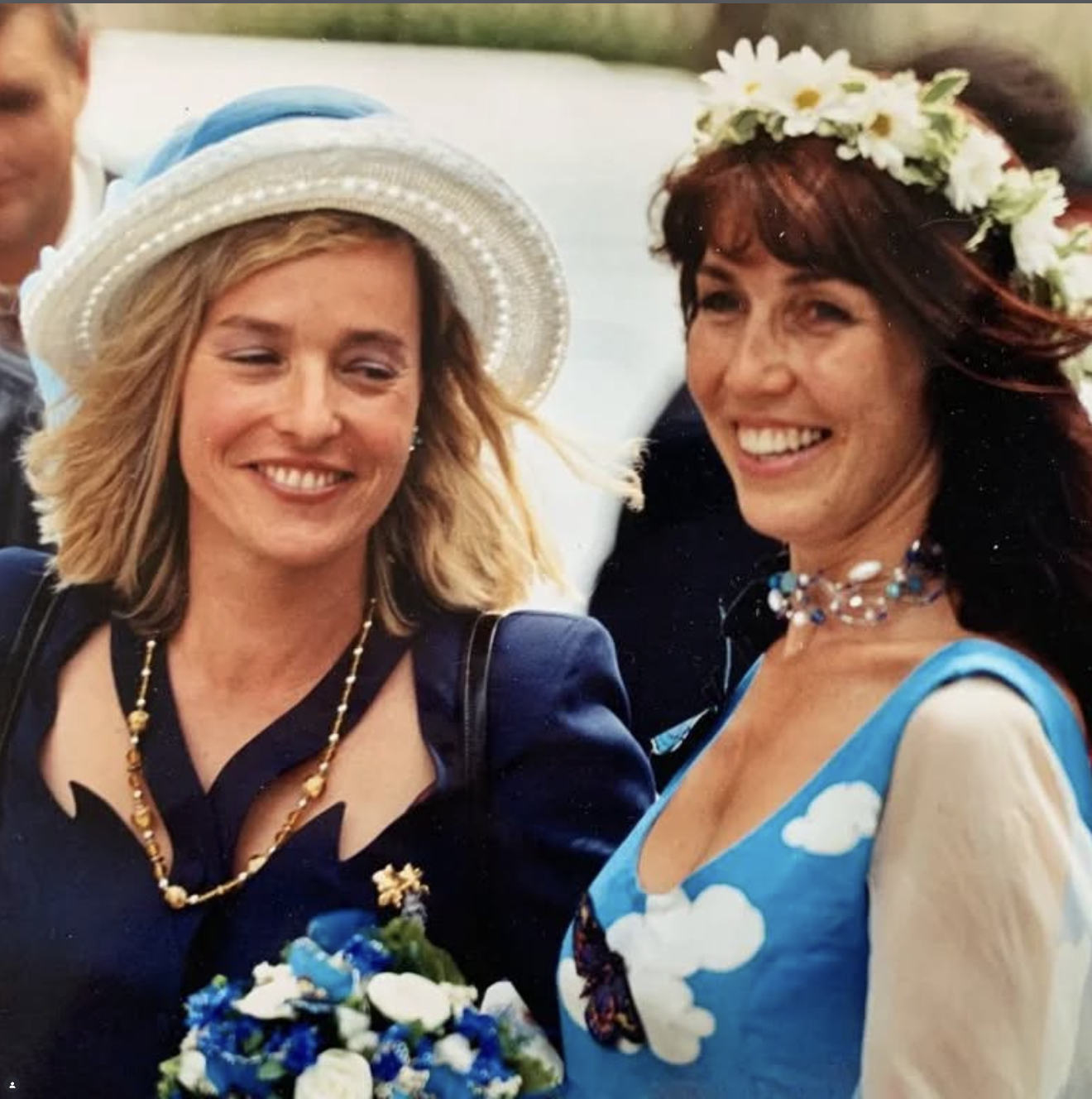 Two women smiling, one holding a bouquet of blue and white flowers. The woman on the left is wearing a large white hat with a blue ribbon, a navy blue outfit, and a gold necklace. The woman on the right is wearing a floral crown, a blue dress with white clouds, and a beaded necklace.