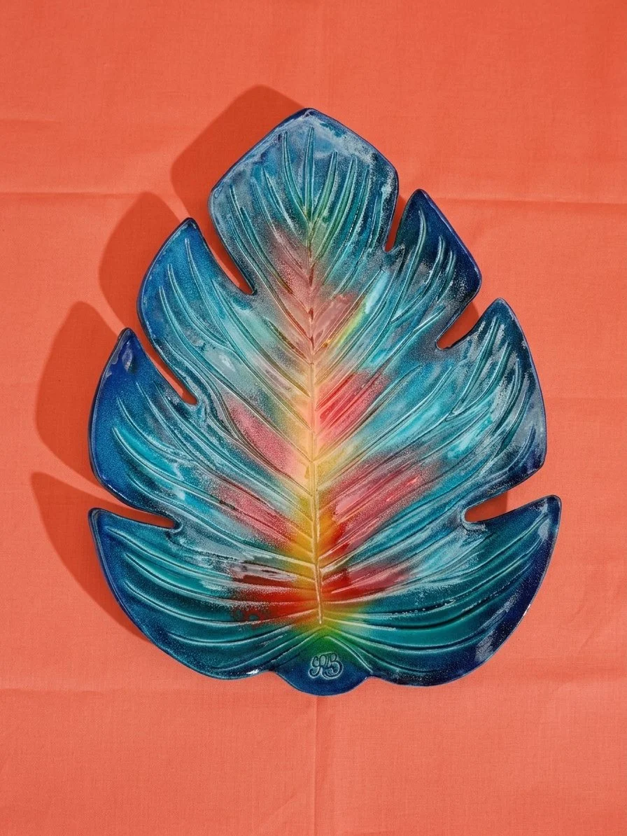 Assiette en forme de feuille tropicale en verre coloré avec dégradés de bleu, vert et rouge sur un fond orange.