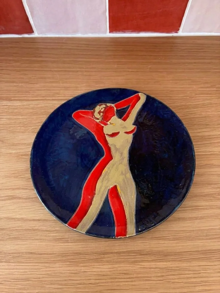Assiette en céramique illustrée d'une femme nue posant avec une main derrière sa tête et l'autre sur ses hanches, sur fond de couleur foncée.