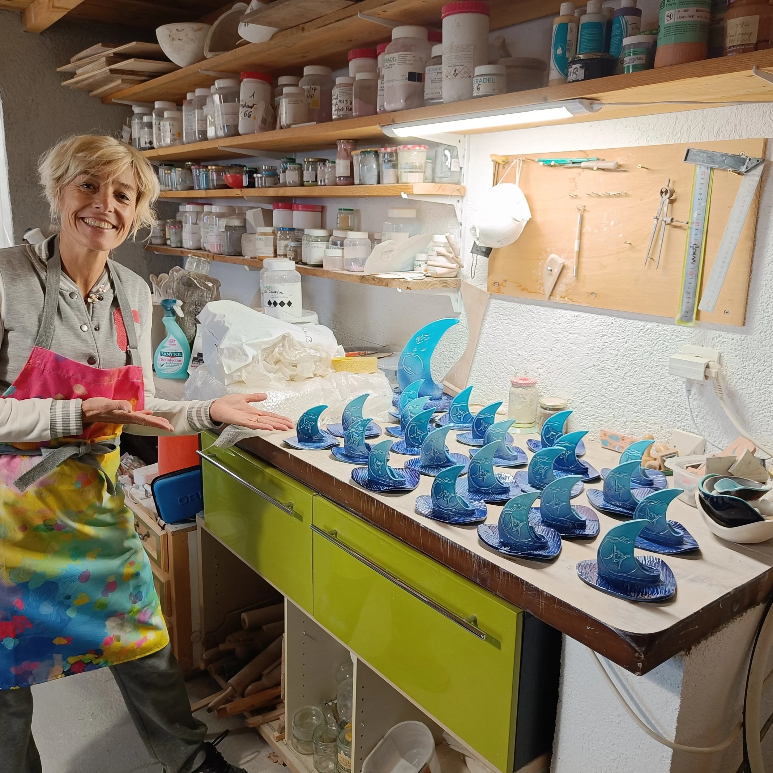 Une personne souriante en artisanat ou atelier, entourée de pots de peinture et outils. Des étagères en bois avec divers contenants et peintures, un plateau avec des objets en verre, et un plan de travail avec des outils de bricolage.