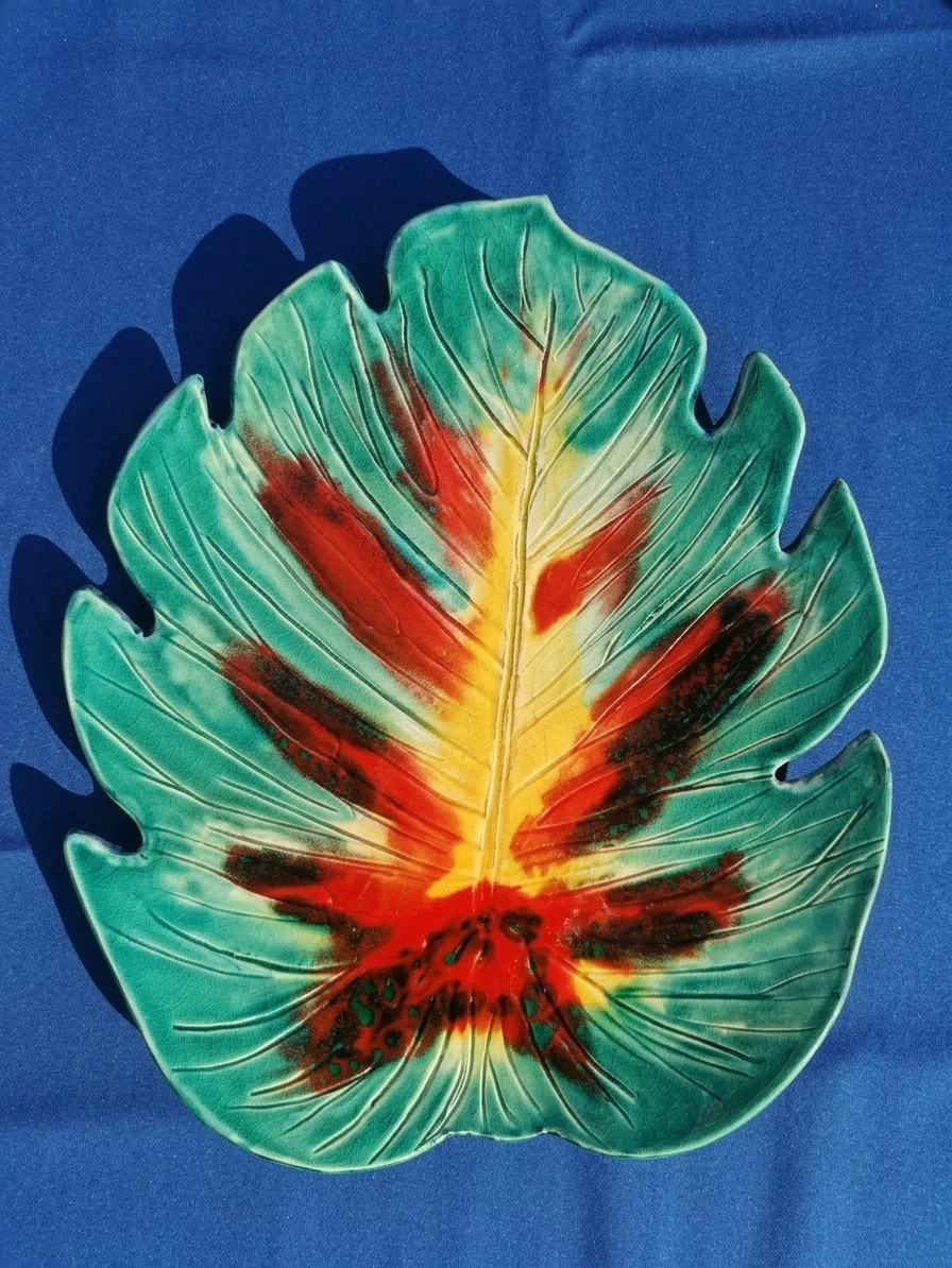 Assiette en forme de feuille végétale avec couleurs vives de vert, jaune, rouge et noir, posée sur un fond bleu.