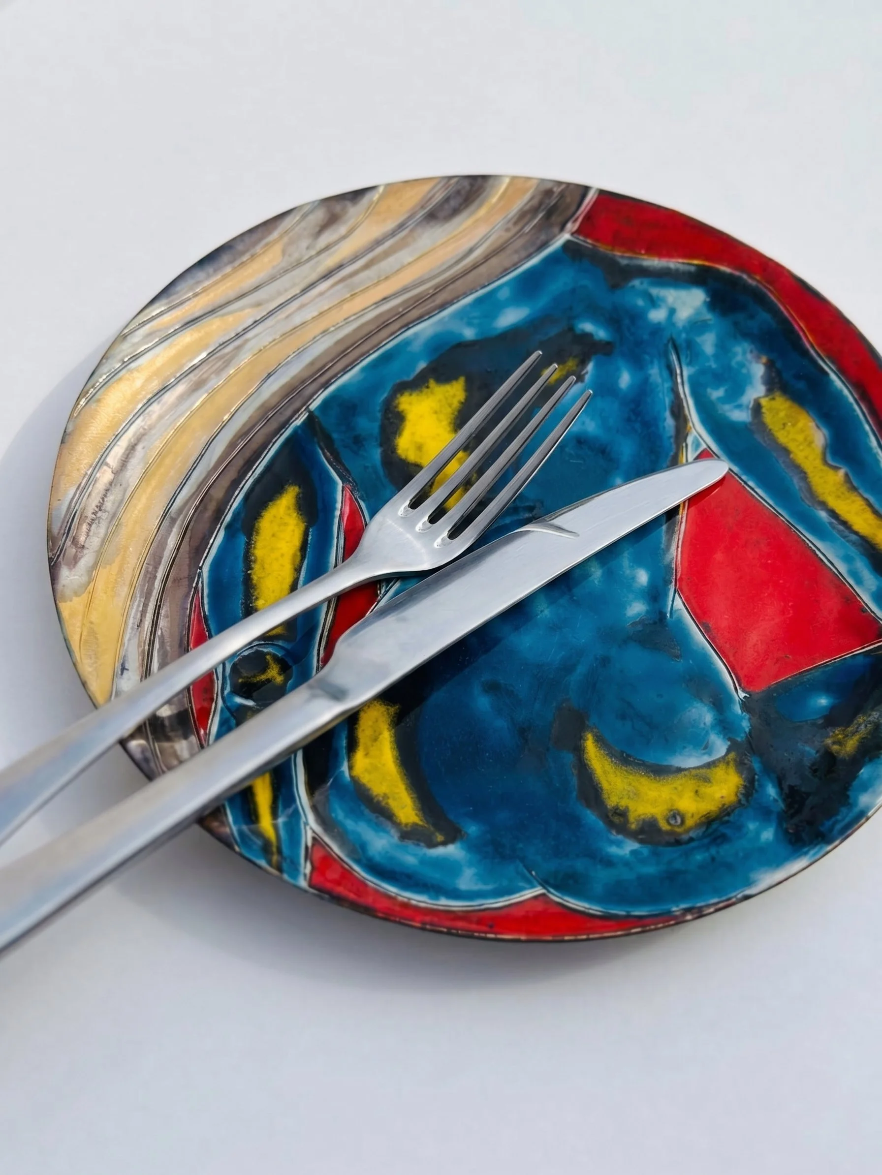 Assiette en céramique colorée avec des motifs abstraits rouges, bleus, jaunes, noirs, accompagnée de trois fourchettes en métal
