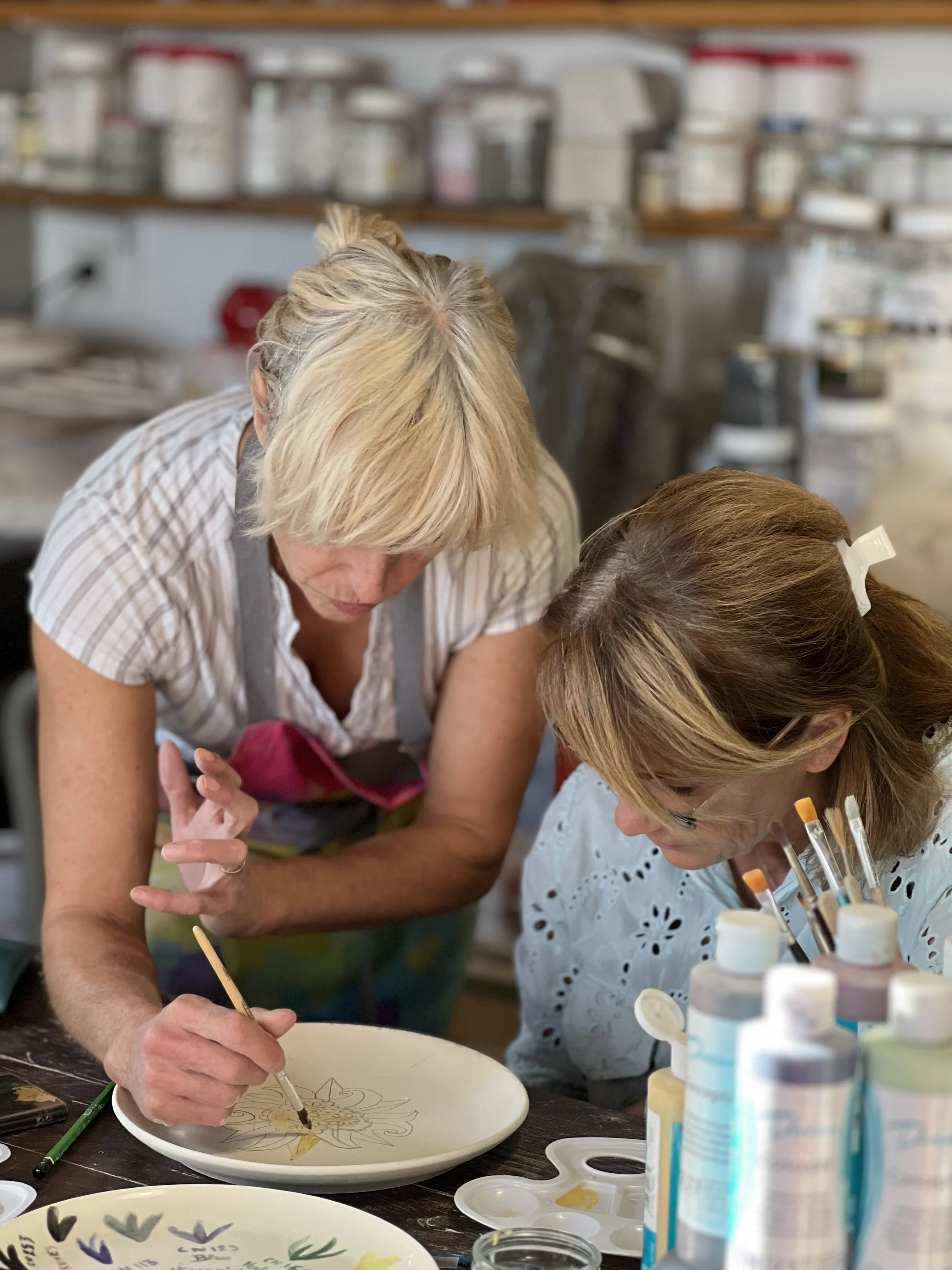 Two women are painting a design on a ceramic plate in an art studio. One woman is using a fine brush, and the other is observing closely. Art supplies and paint bottles are visible on the table.