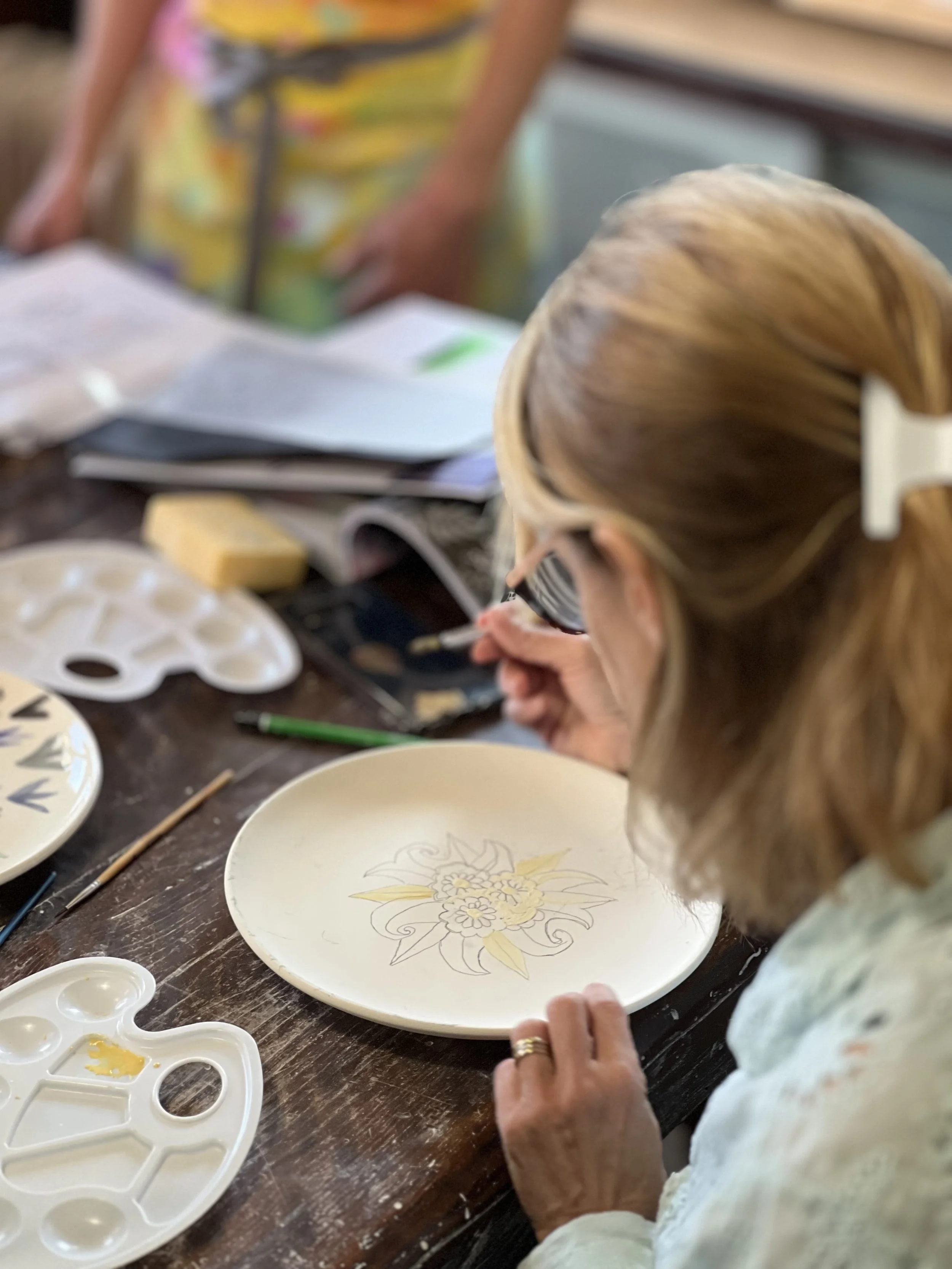 An elderly woman with blonde hair and glasses paints a floral design on a ceramic plate at a craft table. Various painting supplies and palettes are scattered around.