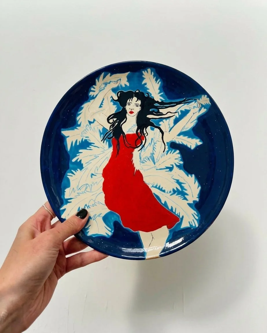 Une assiette décorative avec une illustration d'une femme en robe rouge, flottant devant un fond de large feuilles blanches et un ciel bleu foncé.