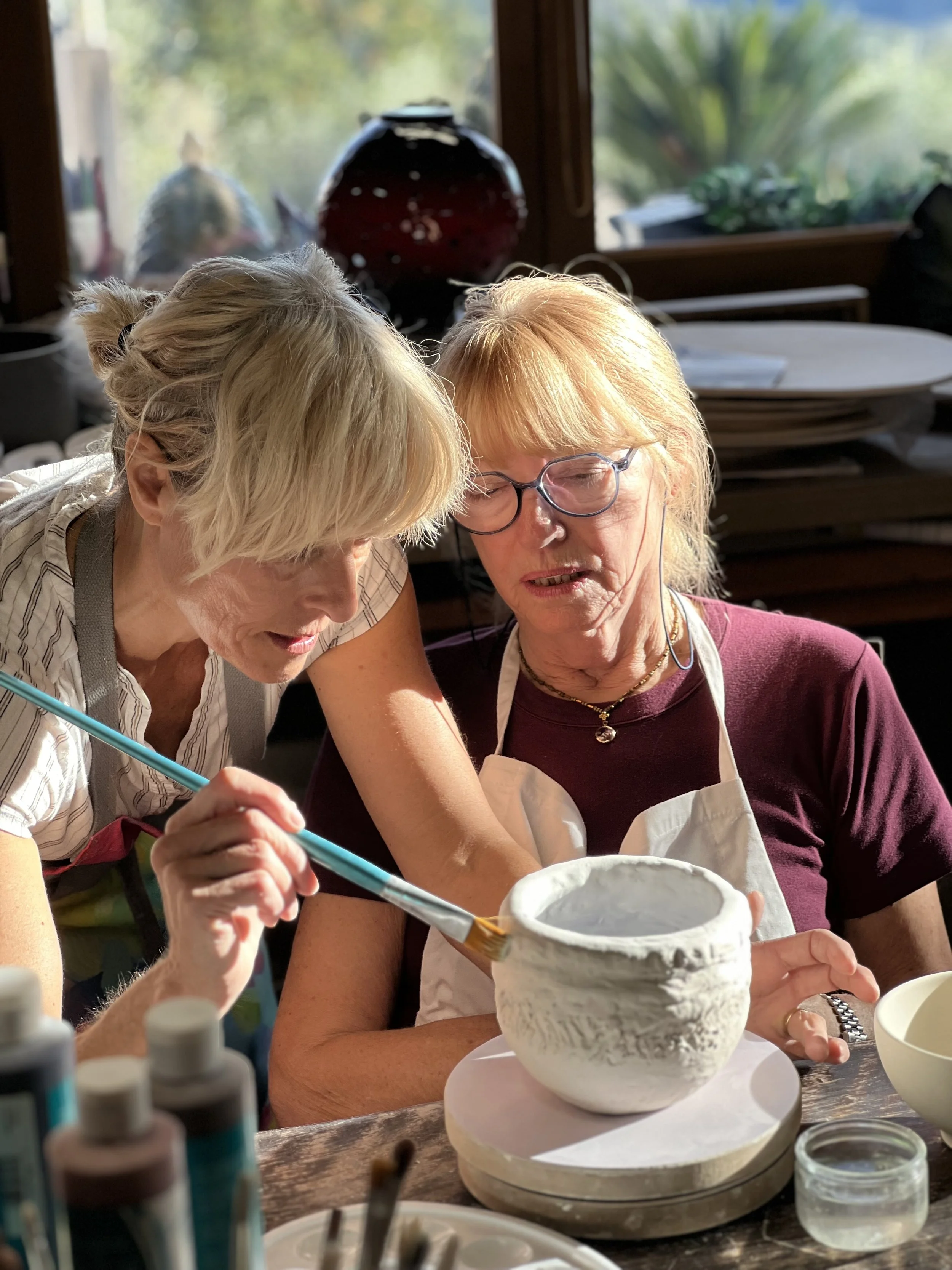 Two women are working together on a ceramic pottery project. One woman is applying glaze with a brush onto a ceramic bowl, while the other woman helps or observes closely. They are seated at a table in a well-lit room with art supplies and pottery in the background.