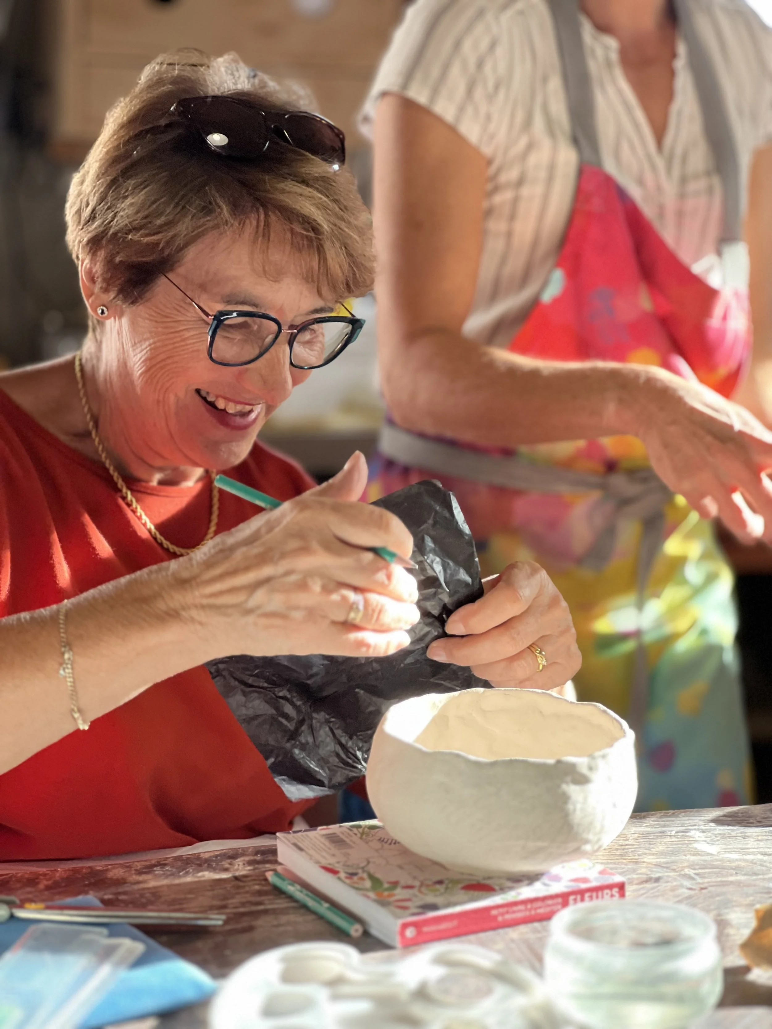 A woman with glasses and earrings, smiling and holding a paintbrush, working on a ceramic piece in a craft setting, with another person in an apron nearby.