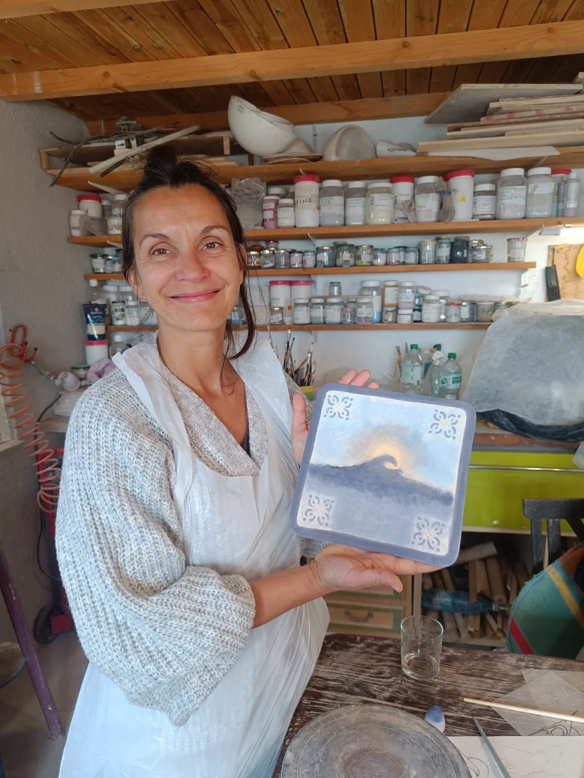 A woman in an art studio holding a small square painting of a wave with the sun setting in the background.