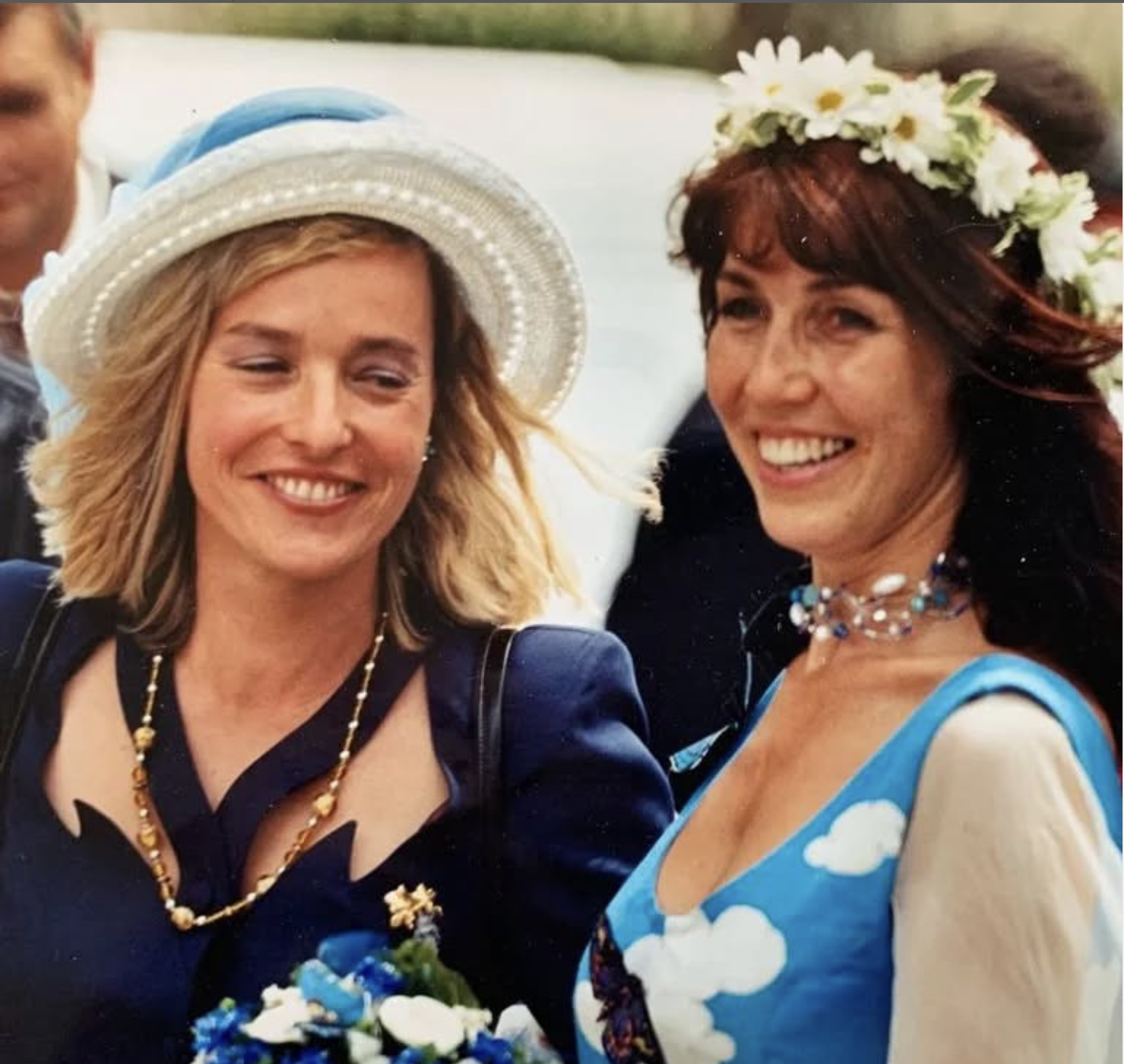 Two women smiling, dressed in colorful attire with floral accessories, outdoors near a body of water.