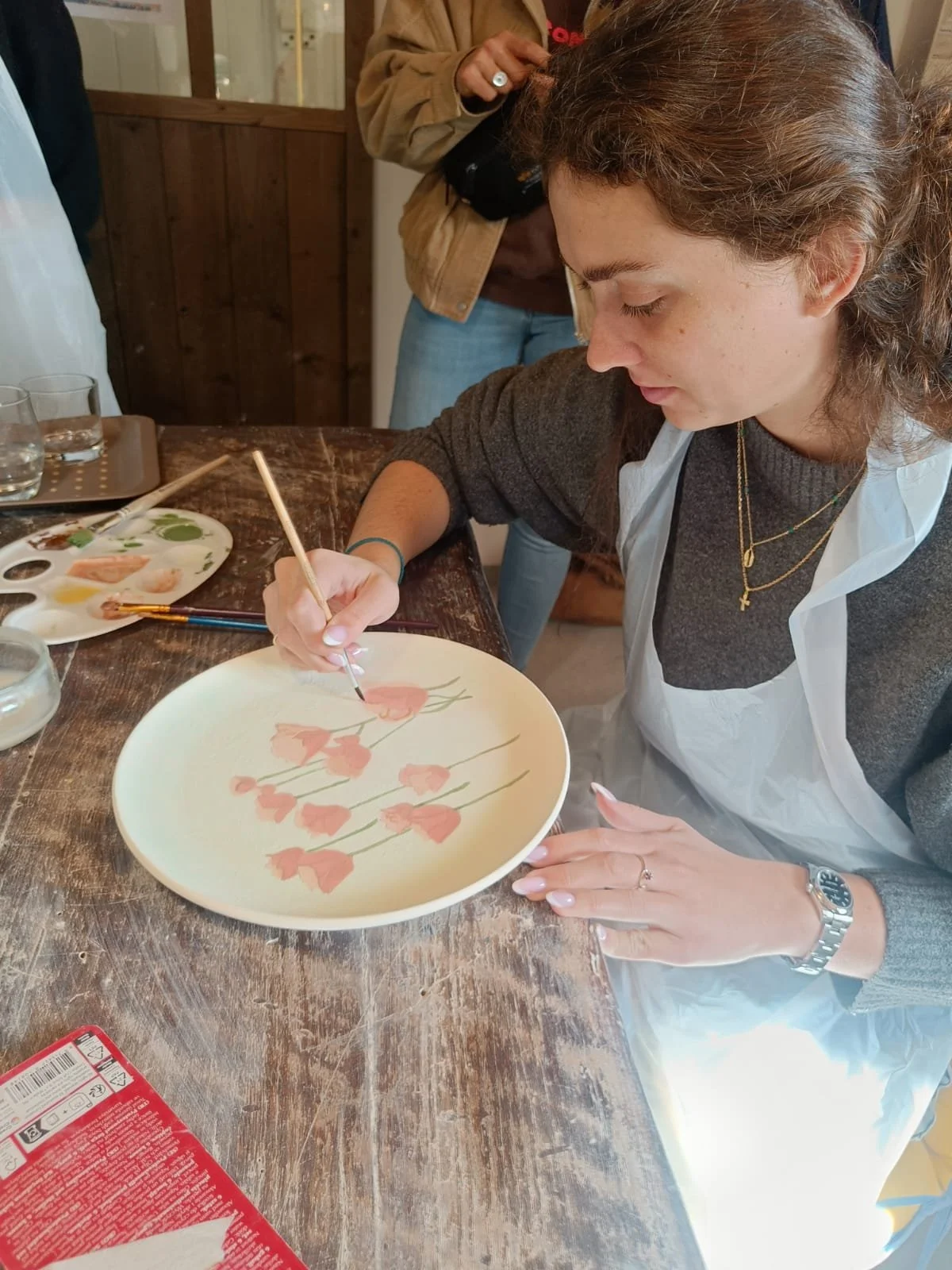 A woman painting a floral design on a ceramic plate with a fine brush, seated at a rustic wooden table with painting supplies around her.