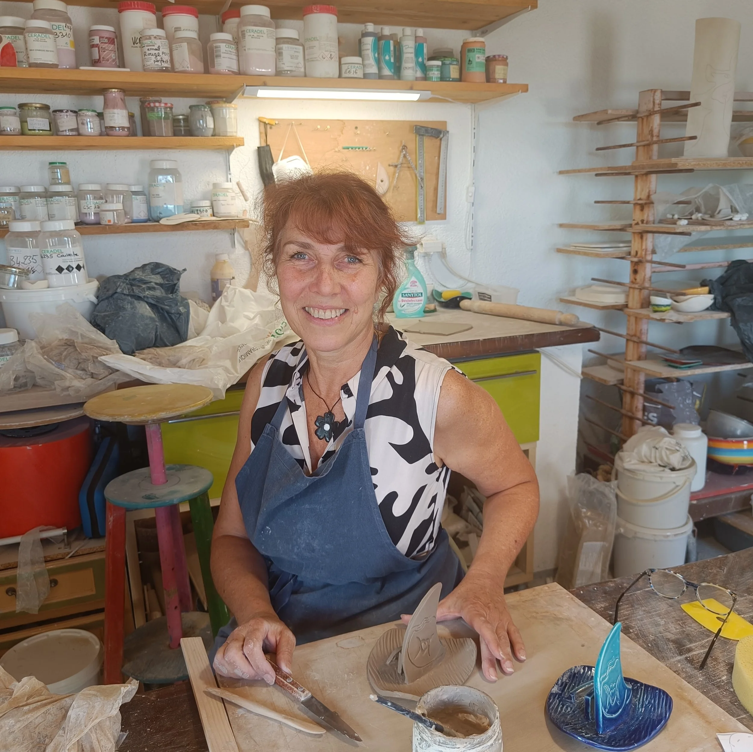 Une femme souriante dans un atelier de poterie, entourée de matériaux et outils pour la sculpture en céramique.