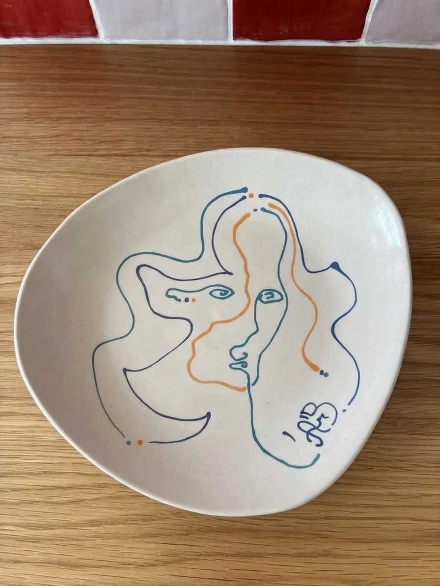 Assiette blanche avec un dessin stylisé en lignes multicolores représentant des visages abstraits et un pentagramme en bas à droite, posée sur une table en bois.
