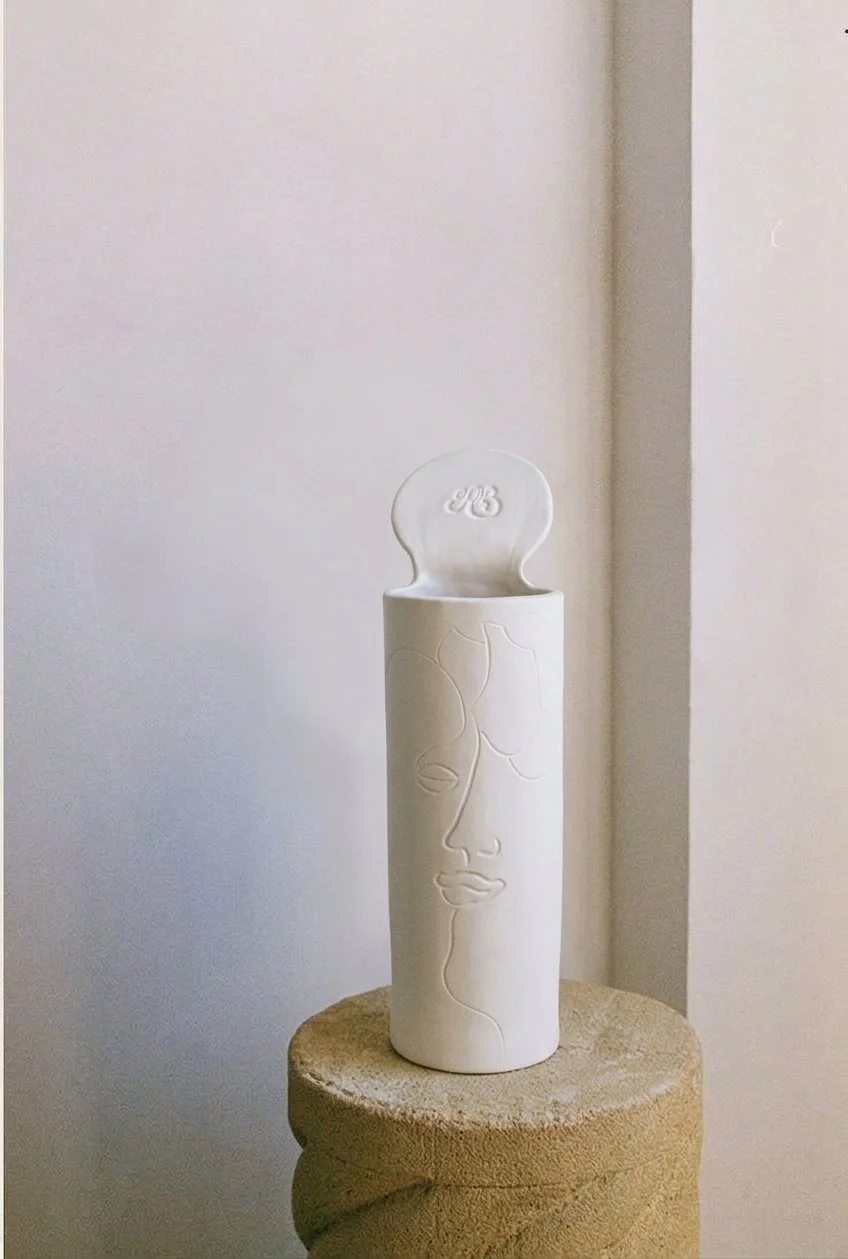 Vase blanc avec un visage stylisé gravé, placé sur un support en bois, dans un intérieur minimaliste.
