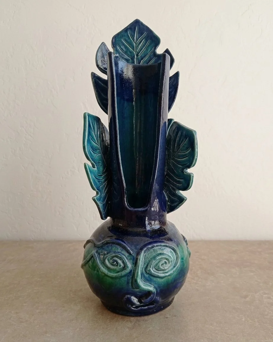 Une vase en céramique avec une décoration de visage stylisé, comprenant des spirales pour les yeux et un nez, et des feuilles en relief qui sortent du haut de la vase, dans des tons de bleu et de vert.