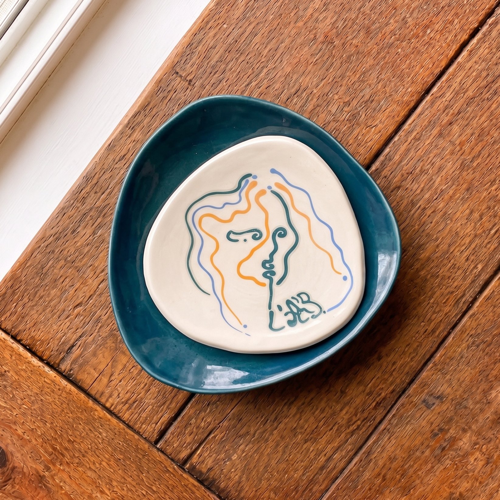 Assiette en céramique avec un dessin abstrait au trait multicolore, posée sur une assiette bleu foncé, reposant sur une table en bois.