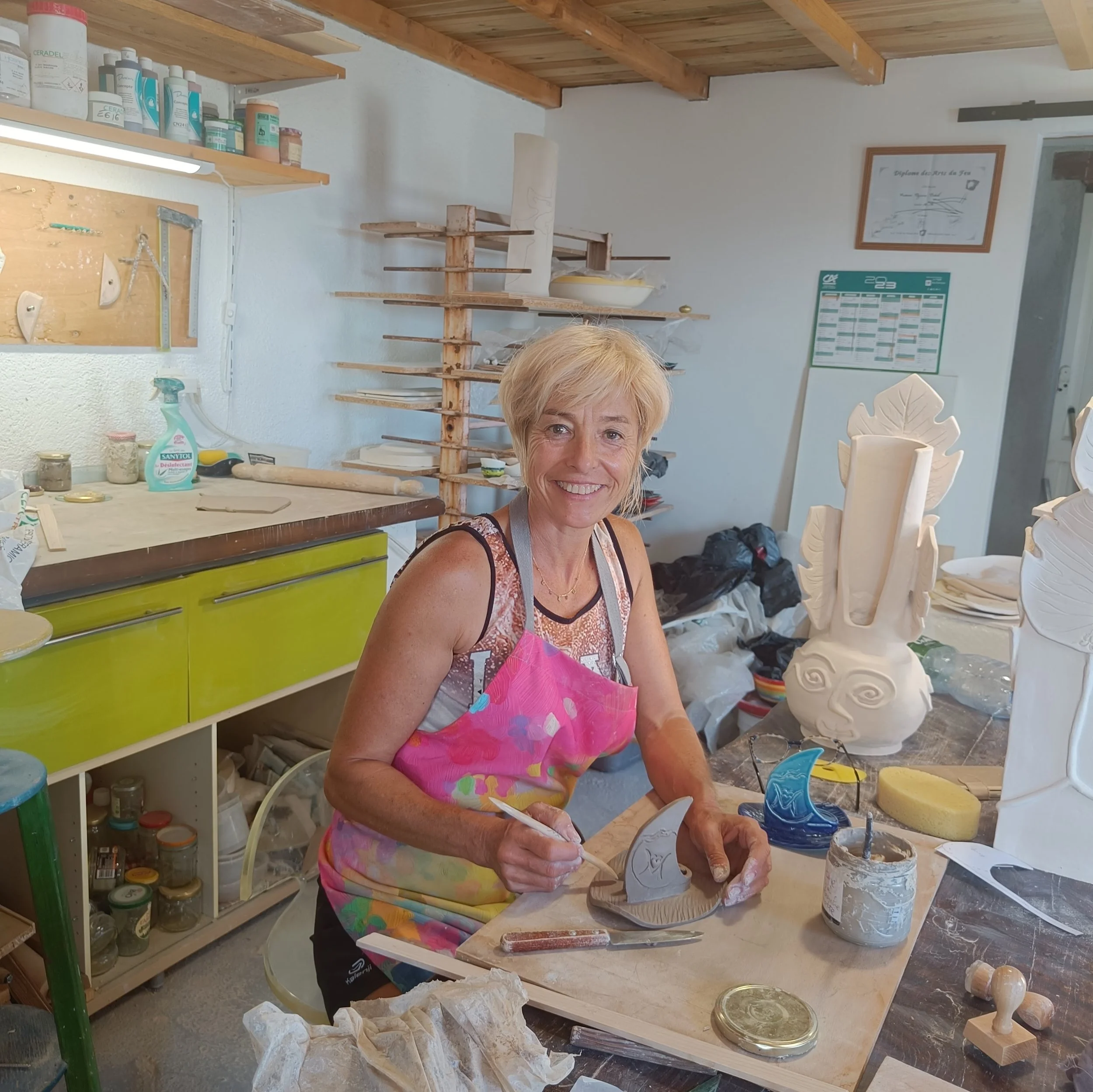 Une femme souriante portant un tablier rose travaille avec de la porcelaine dans un atelier de céramique. Elle conçoit une sculpture de tête de sirène en argile. L'atelier est rempli d'outils, de pots de peinture, et d'objets en céramique en cours de
