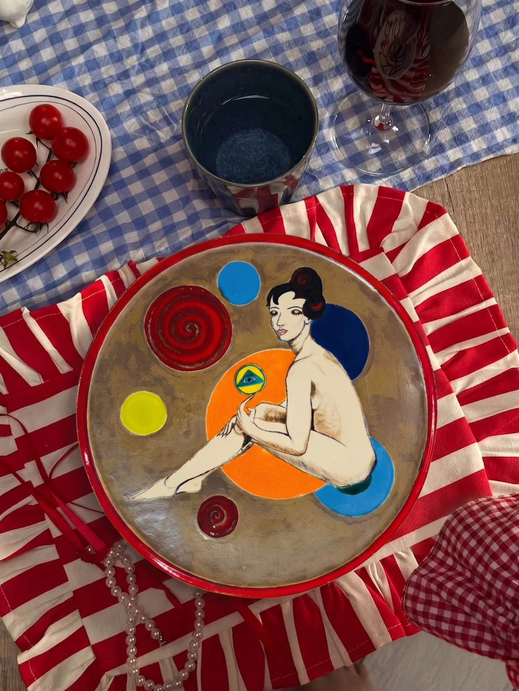 Assiette avec une illustration artistique d'une femme nue assise, entourée de cercles colorés, placée sur une nappe à carreaux rouge et blanc. À côté, un plateau avec des tomates cerises, un verre de vin rouge, un verre vide, et un collier de perles