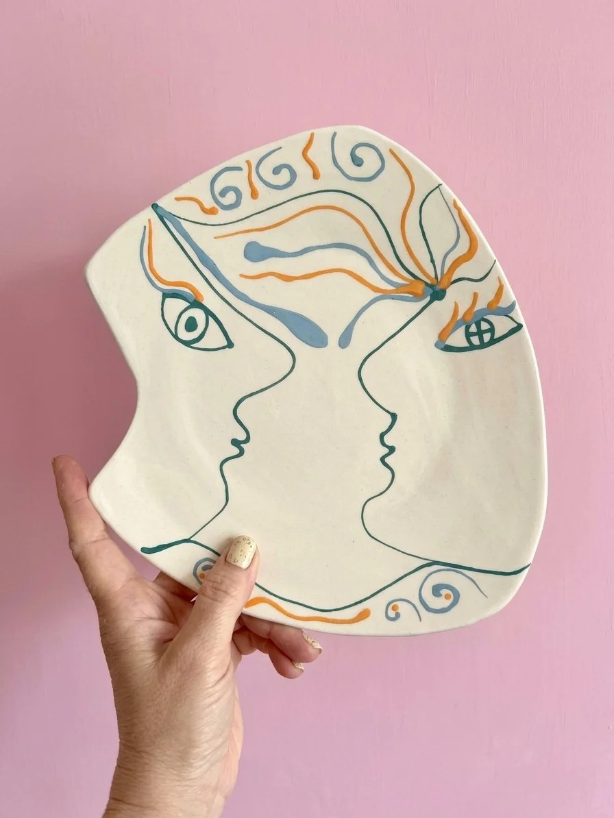 Une assiette en céramique avec un dessin abstrait de deux visages qui se font face, peint avec des lignes noires, orange et bleues, étant tenu par une main contre un mur rose.