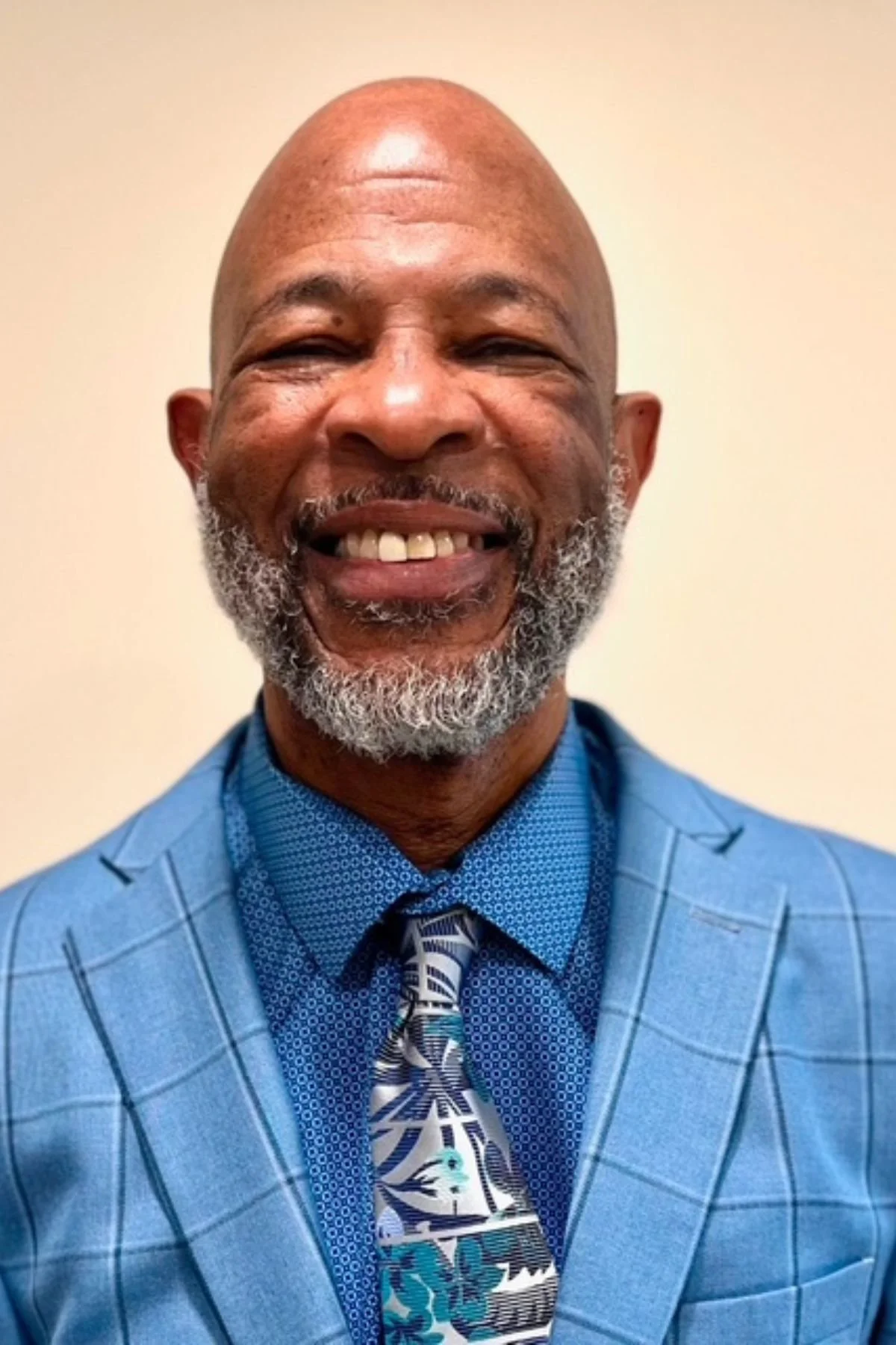 A smiling African American man with a gray beard, wearing a blue checkered suit, a blue patterned shirt, and a colorful abstract tie, standing against a plain background.