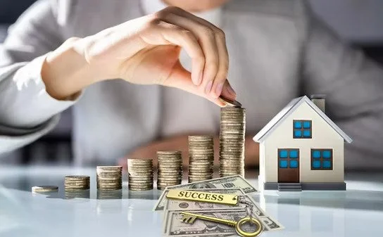 Hand stacking coins next to a small model house and keys on a table, representing real estate investment and financial growth.