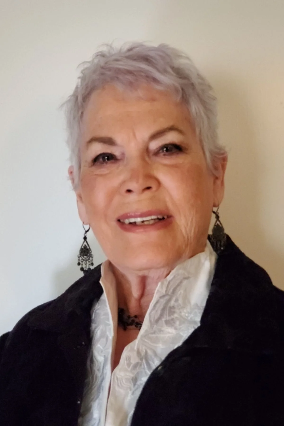 A smiling older woman with short gray hair, wearing a white shirt, black jacket, and black earrings, against a plain background.