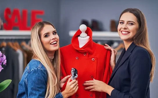 Two women shopping in a clothing store, holding a red coat on a mannequin and smiling at the camera.