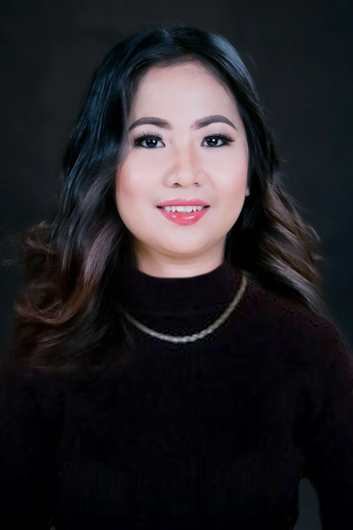 A woman with long, wavy black and brown hair smiling, wearing a black top and a pearl necklace, against a dark background.