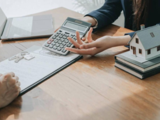 Two people conducting a real estate transaction, reviewing documents, using a calculator, with a miniature house model on a stack of books on a wooden table.