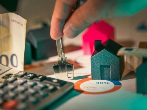Close-up of a person's hand placing a key into a crucial-looking lock, with small house models, a calculator, and a pie chart on a desk, suggesting real estate or financial planning.