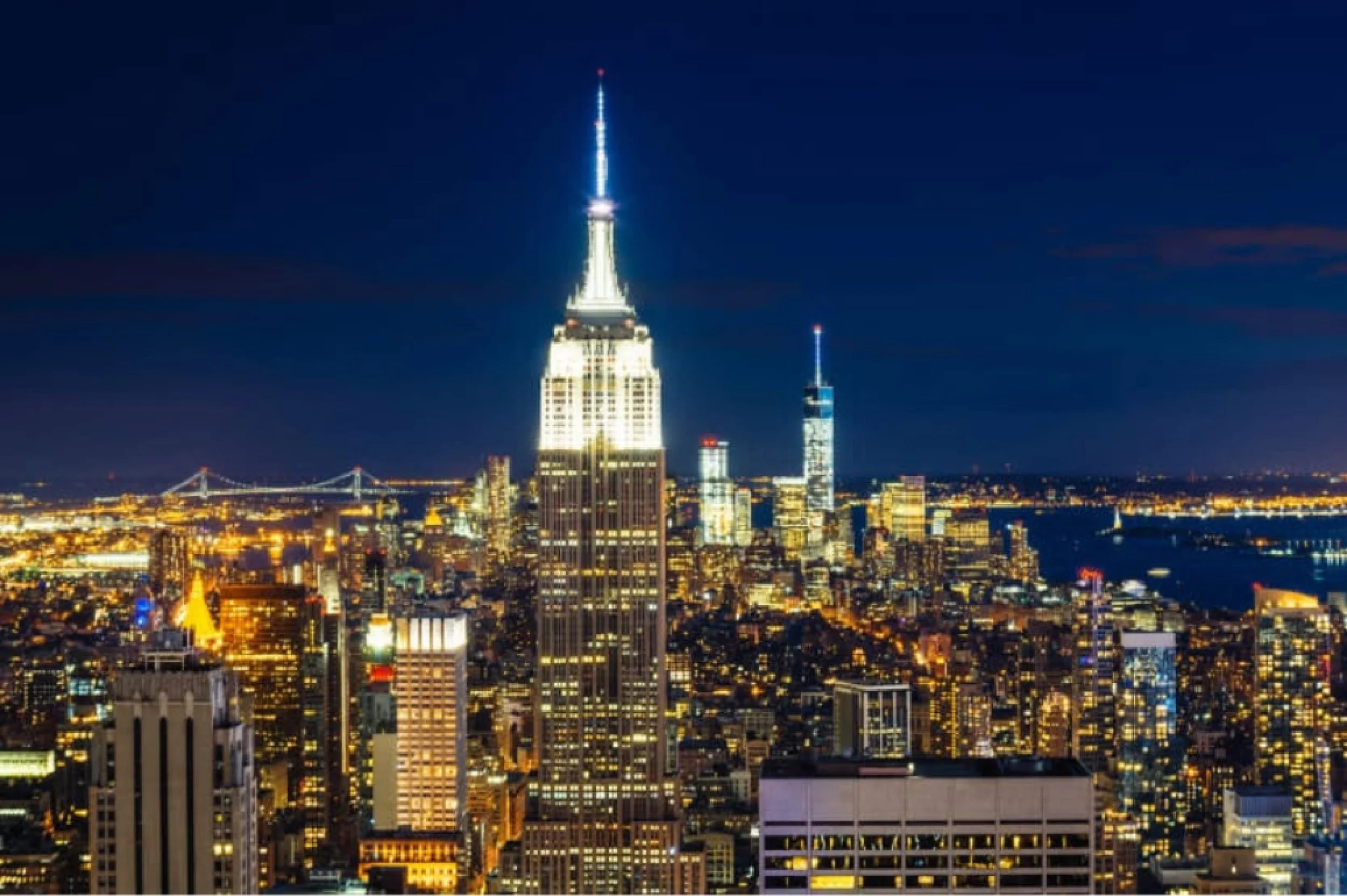 Nighttime cityscape of New York City featuring the Empire State Building illuminated and the One World Trade Center in the distance.