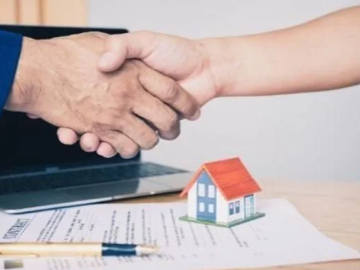 Two people shaking hands on a desk with documents, a pen, a small model house, and a laptop.