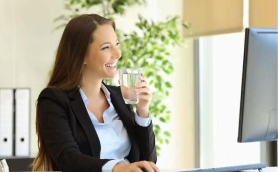 A woman in business attire smiling and drinking a glass of water while working at her desk in an office.