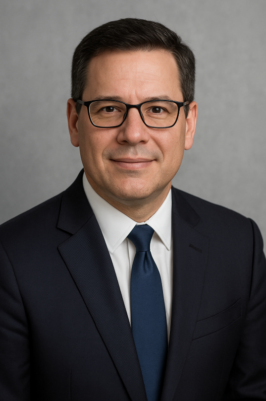 Headshot of a man wearing glasses, a dark suit, white shirt, and blue tie, against a gray background.