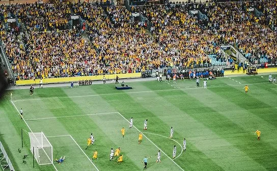 A soccer match in a large stadium with players on the field, referees, and a crowd of spectators in the stands.