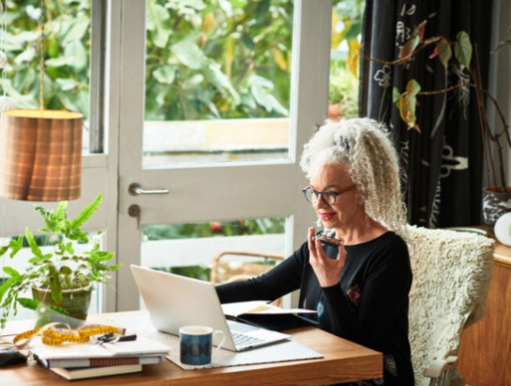 An elderly woman with gray curly hair and glasses sitting at a wooden desk indoors, talking on a mobile phone, with a laptop, a mug, notebooks, and plants nearby, and a window with greenery outside.