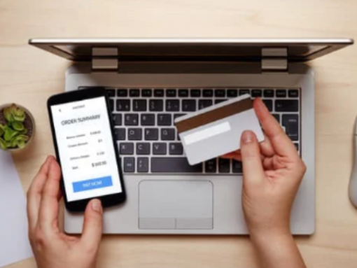 Person holding a credit card and a smartphone displaying an order summary, with a laptop open on a light-colored desk.