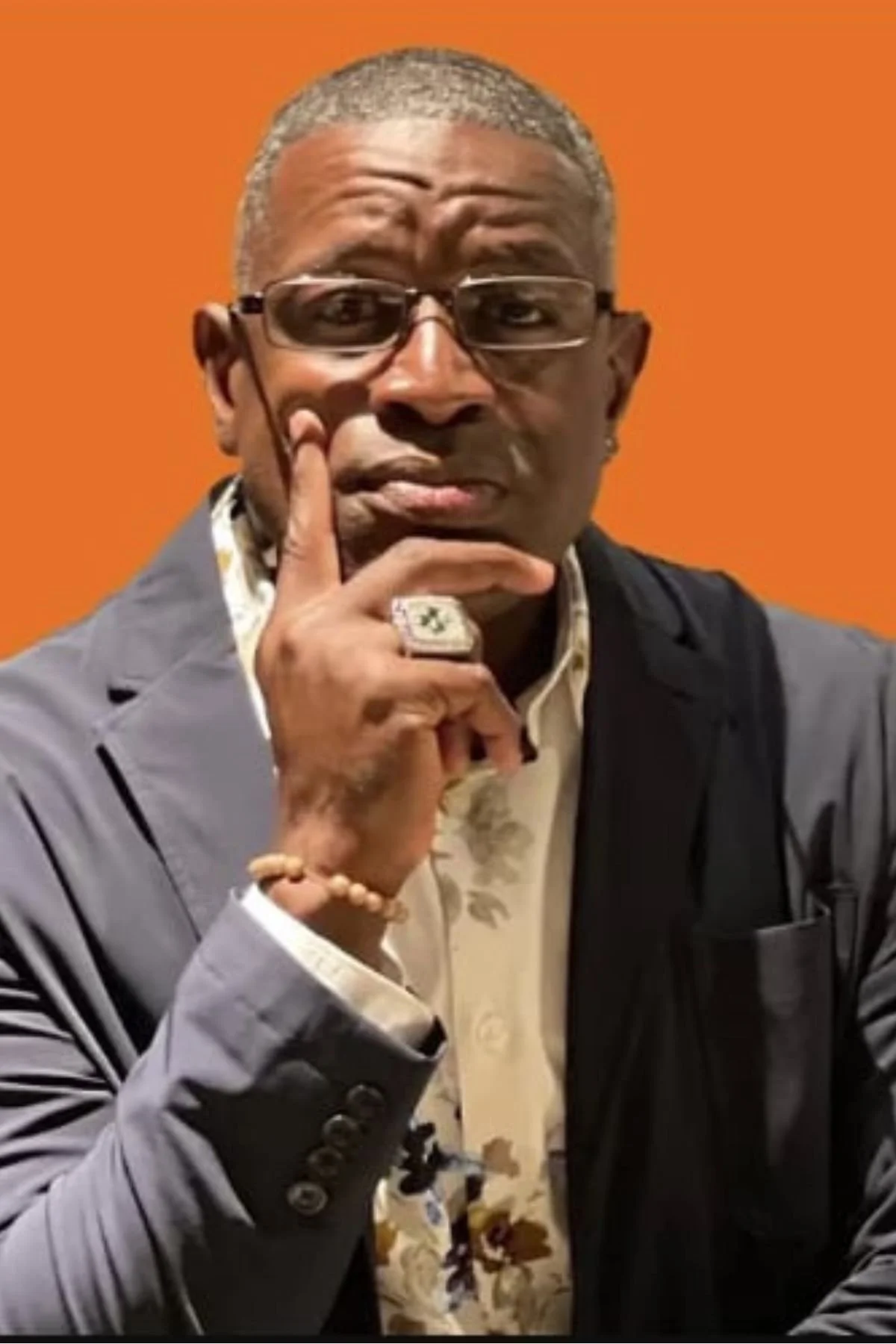 A man with short gray hair, glasses, and dark skin, wearing a patterned shirt and a gray blazer, is posing with his hand on his chin against an orange background.