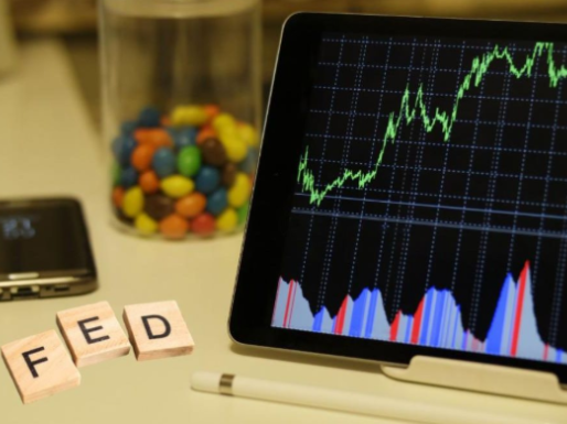 A tablet displaying a financial stock chart on a desk with colorful candies in a jar, Scrabble tiles spelling 'FED,' a pen, and a smartphone in the background.