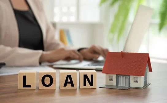 Wooden blocks spelling 'LOAN' placed on a desk, next to a small model house. A person in business attire is working on a laptop in the background, with greenery and a window behind.