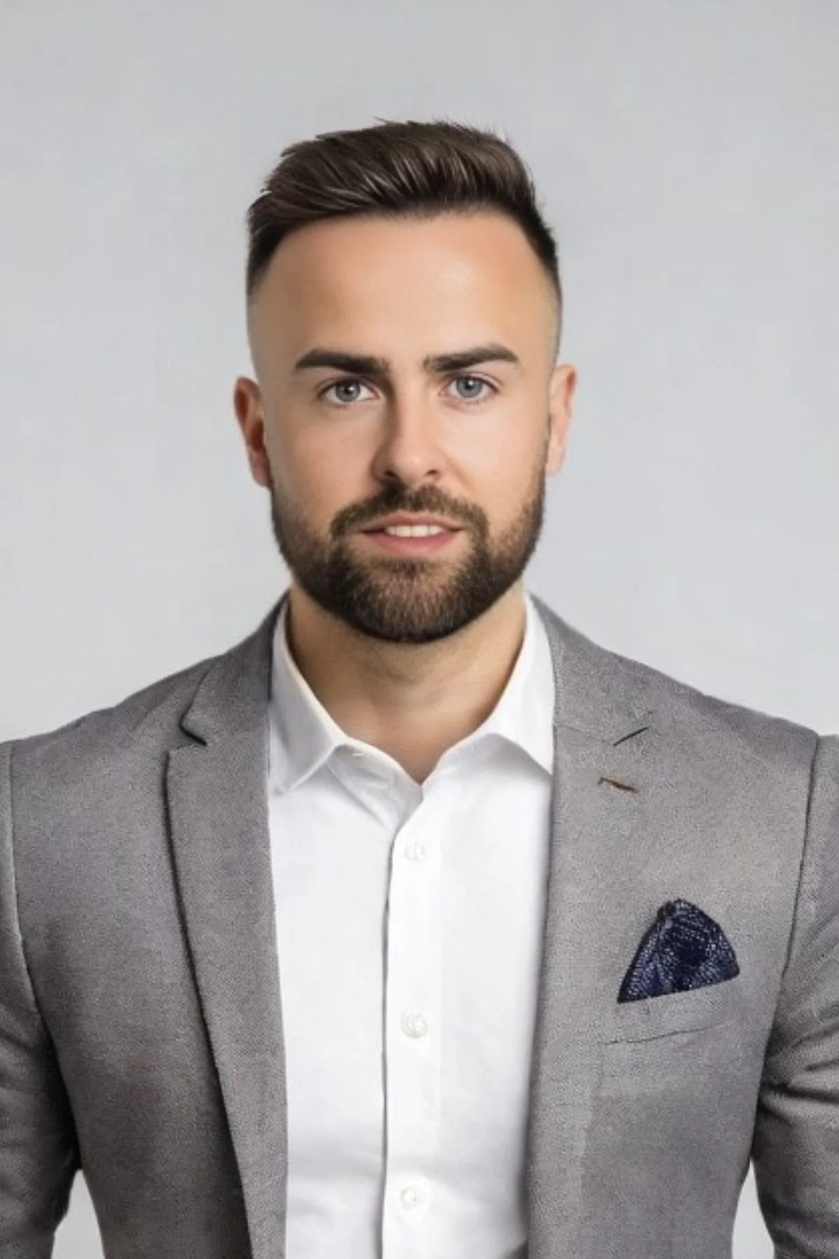 A well-dressed man with a beard and styled hair wearing a grey suit jacket, white shirt, and a dark pocket square, standing against a plain light background.