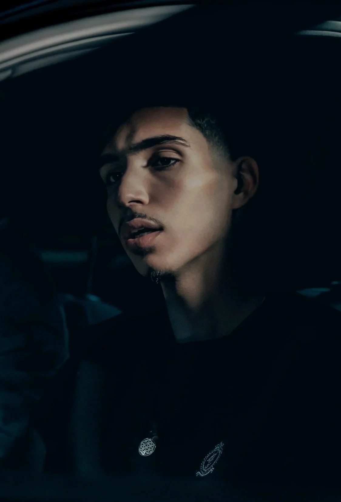 A young man with short hair and a mustache looking at the camera in a dark setting, with a slight smile, wearing a black shirt with a small emblem near the collar.
