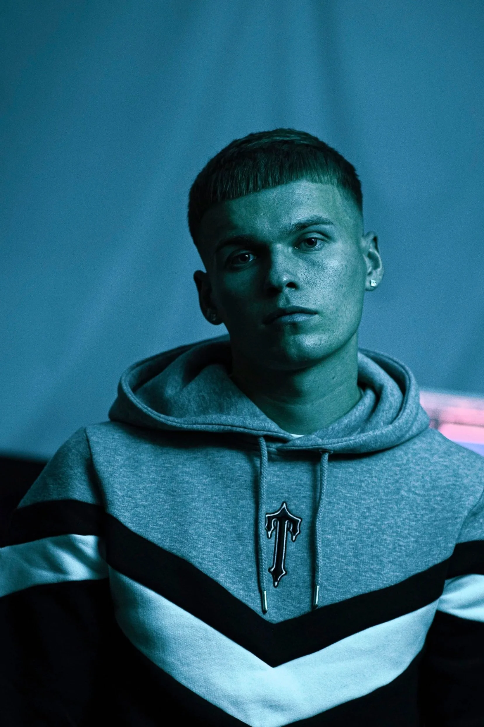 A young man with short hair and earrings, wearing a gray hoodie with black and white stripes and a logo on the front, looking directly at the camera in a blue-lit room.