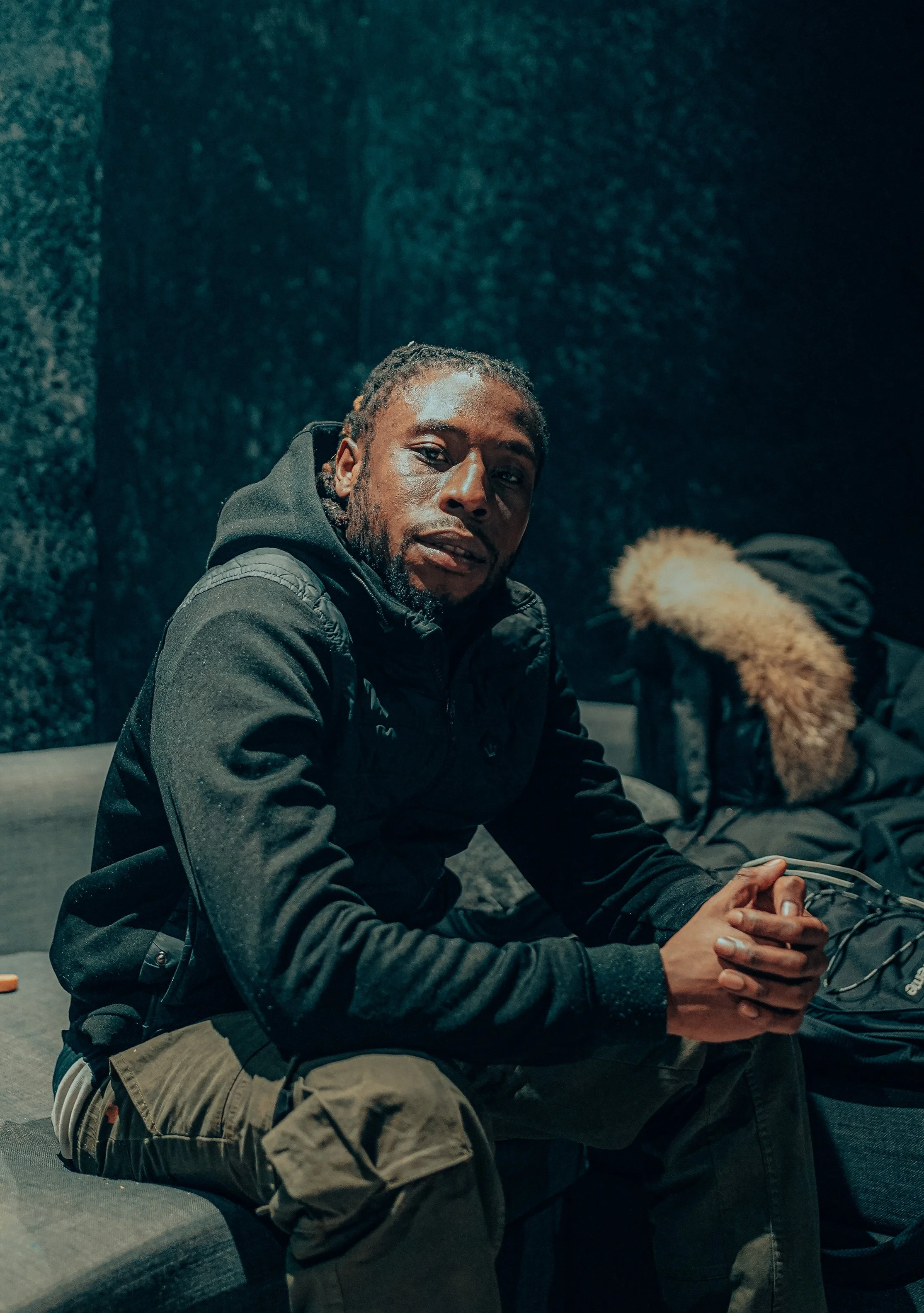 A man with dreadlocks wearing a black hoodie and camouflage pants sitting on a gray couch in a dimly lit room, looking at the camera.