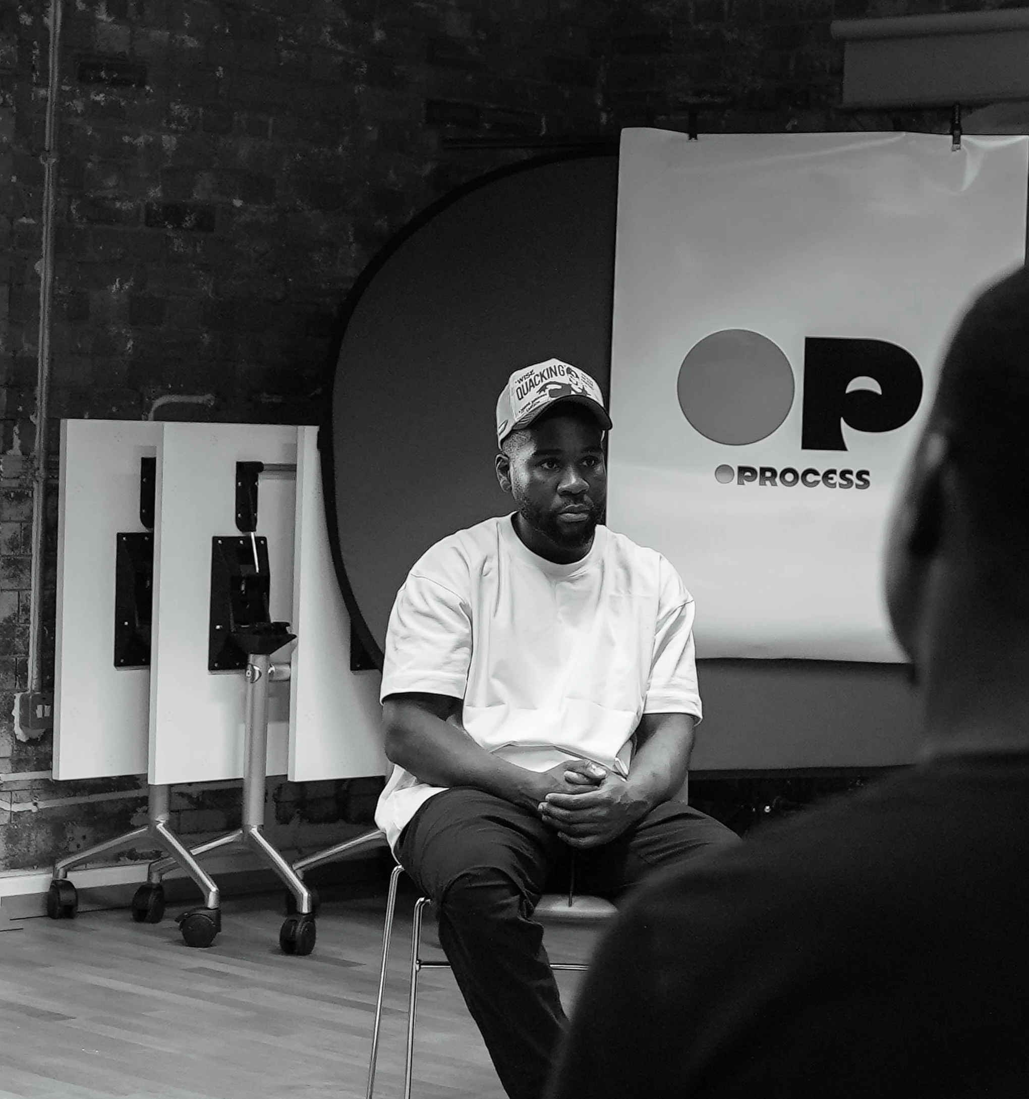 A man wearing a cap and a white t-shirt is sitting on a chair during a discussion or interview in a modern room with a brick wall and a branded backdrop.