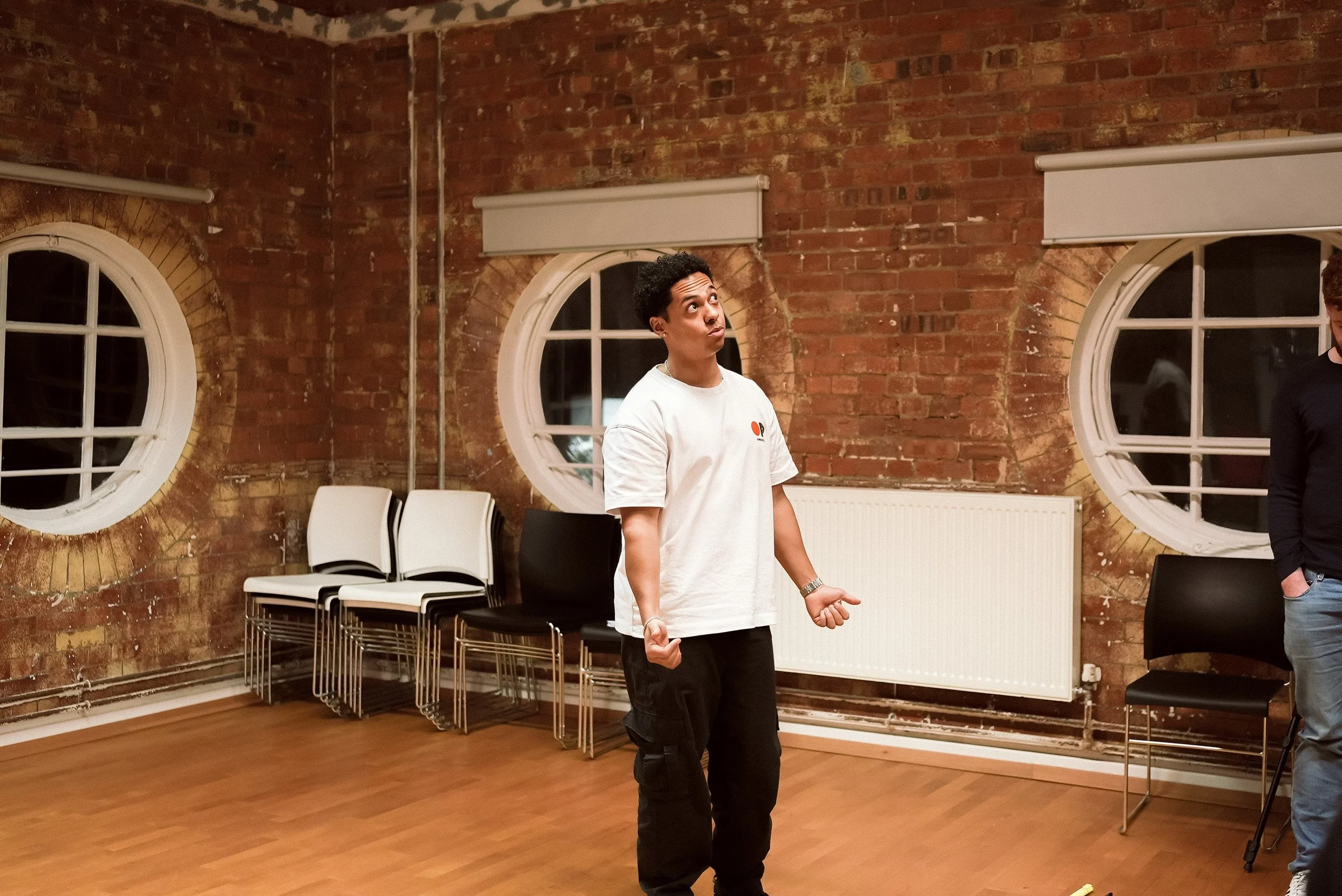 A young man standing in a room with exposed brick walls and round windows, gesturing with his hands, wearing a white T-shirt and black pants, with chairs stacked against the wall behind him.