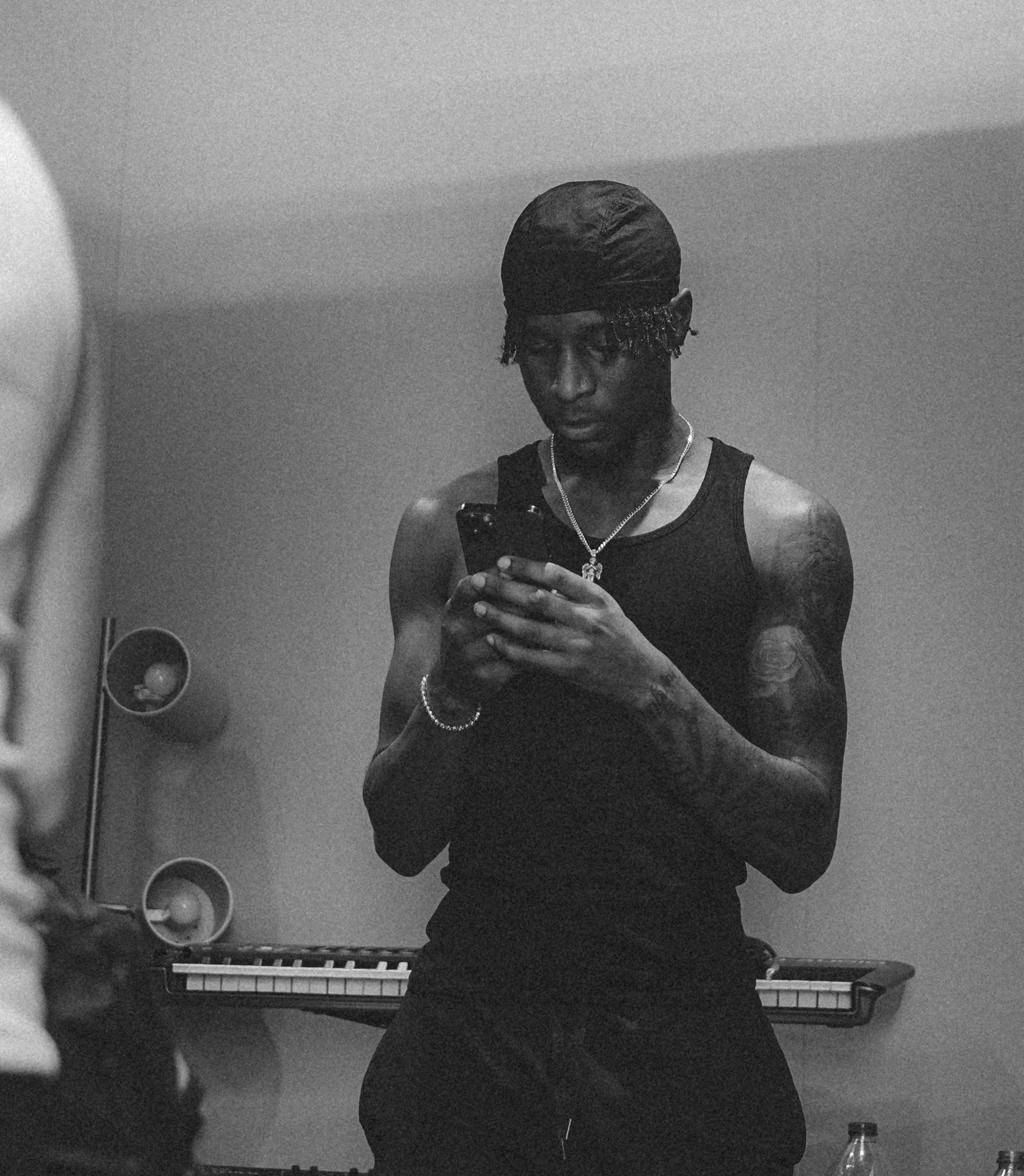 A young man in a black tank top, wearing a bandana and jewelry, takes a mirror selfie in a room with a keyboard and speakers in the background.