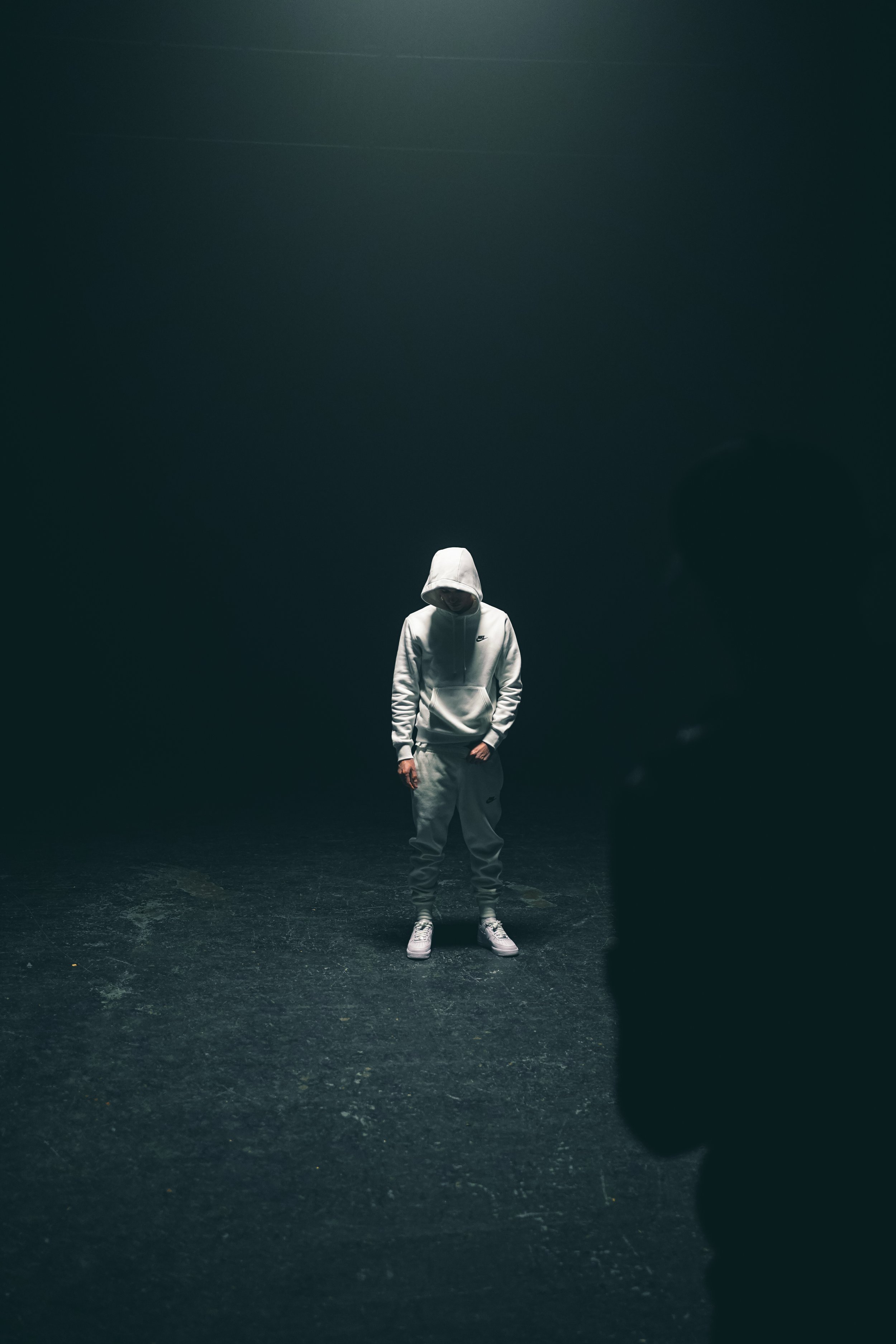 A person dressed in a white hoodie and sweatpants stands alone in a dark, empty space, with a dark shadowy figure partially visible in the foreground.