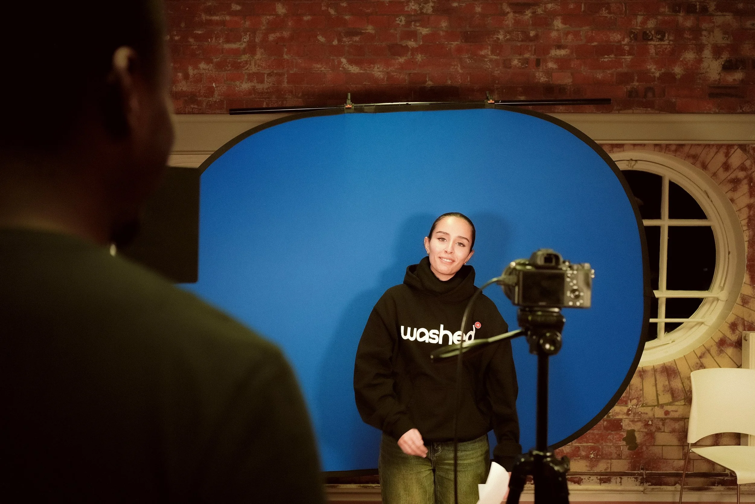 Woman in black hoodie with white text standing in front of blue screen during a photo shoot, man with camera capturing her, brick wall and a window in the background.