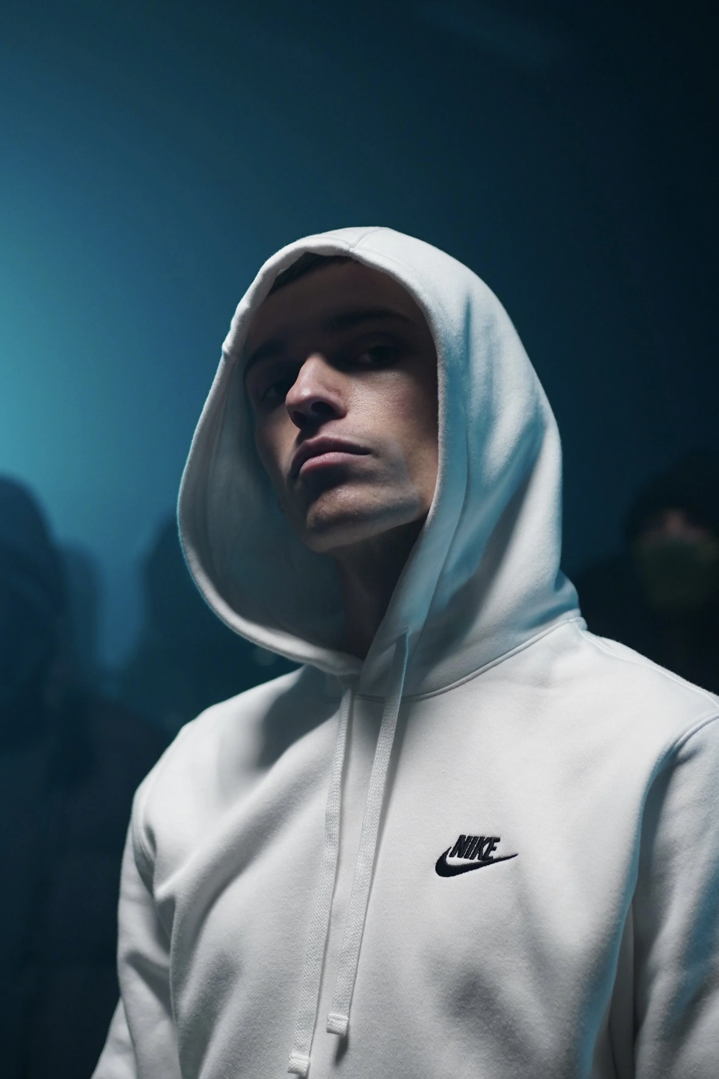 A young man wearing a white Nike hoodie with the hood up, looking into the camera with a serious expression, in a dimly lit environment.